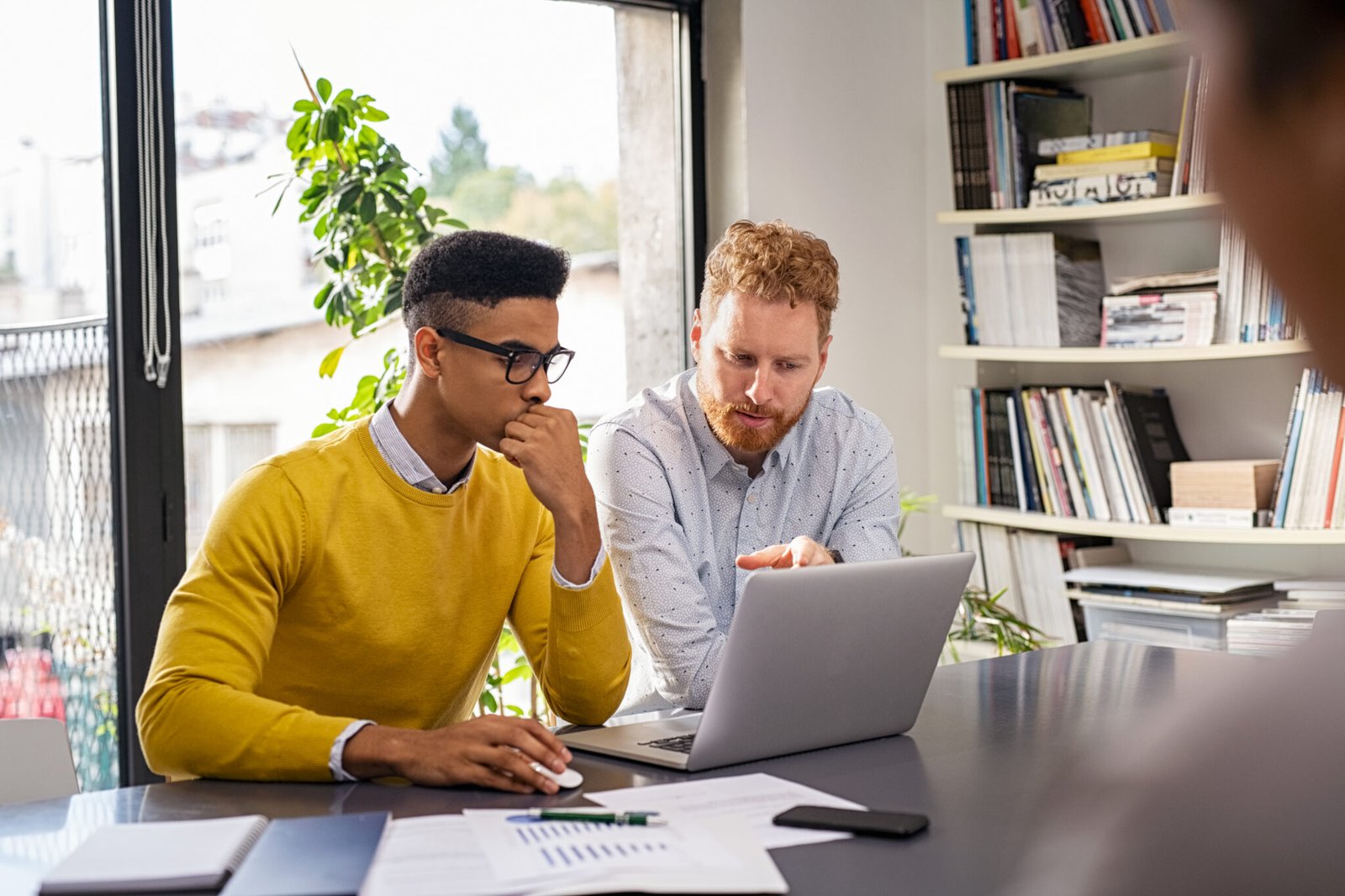 Mature and young businessmen discussing during meeting in boardroom. Business executive working with apprentice in creative office. Multiethnic business team in smart casual with partner working together on laptop.